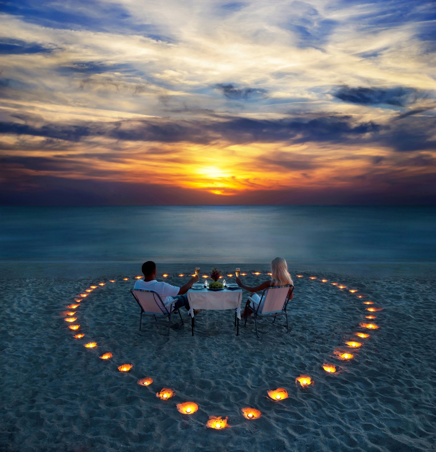 A couple sitting at a table on a beach with candles in the shape of a heart

Description automatically generated