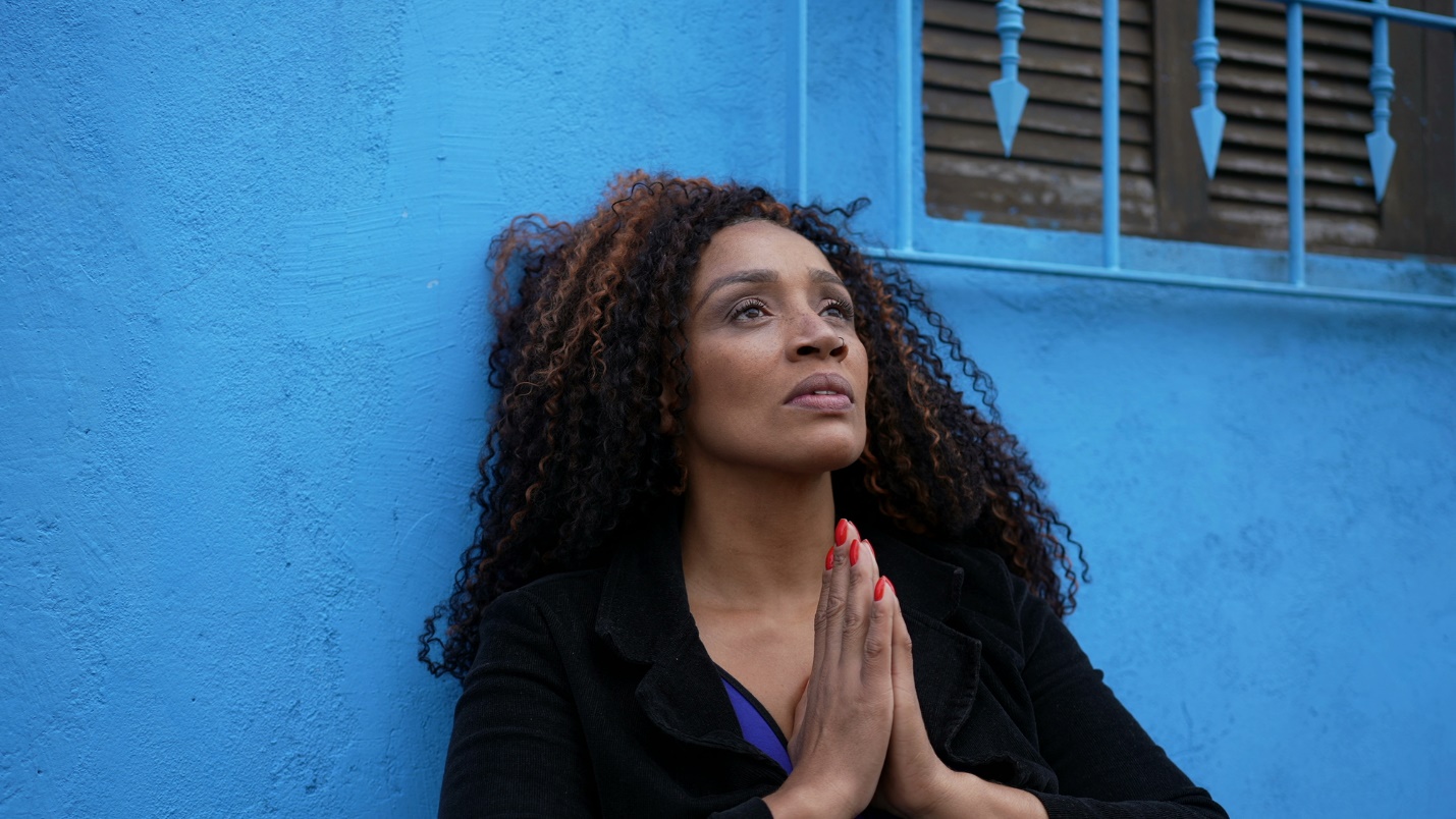 A person with curly hair and hands together in front of a blue wall

Description automatically generated